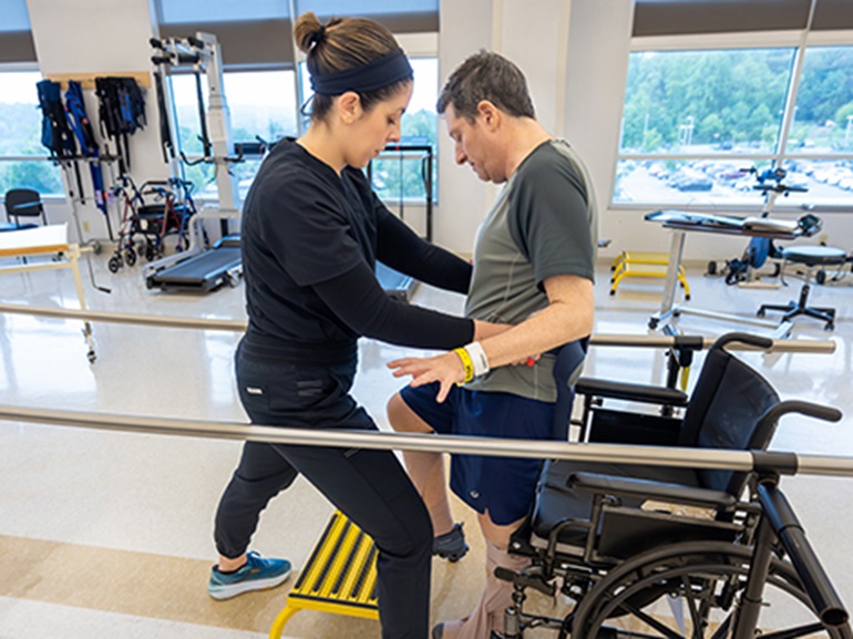 A male patient works to stand from his wheelchair and practice walking again after brain injury with the help of his therapist.