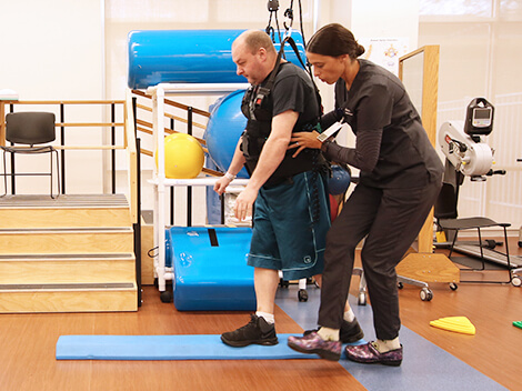 A male patient walks in the therapy gym with the support of his therapist and an overhead, weight supportive harness.