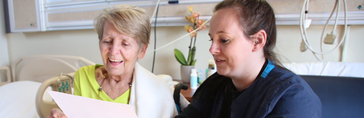 A female nurse reviews admission information with a patient sitting beside her hospital bed.