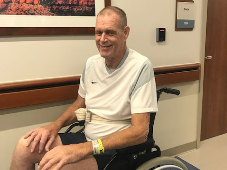 Bruce sits in his wheelchair in the hospital hallway wearing a white t-shirt.