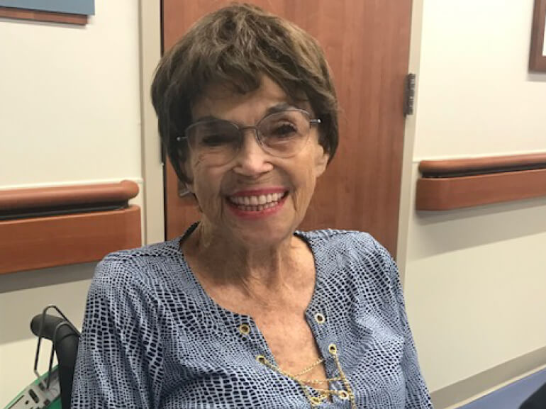 Sylvia sits in her wheelchair in the hospital hallway, smiling at the camera.