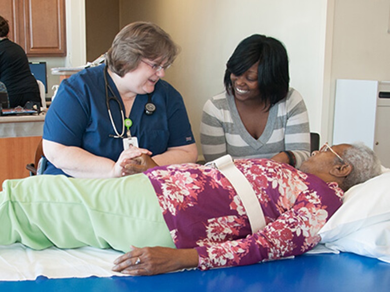 A female nurse holds their patient's hand while the patient lays beside their loved one.