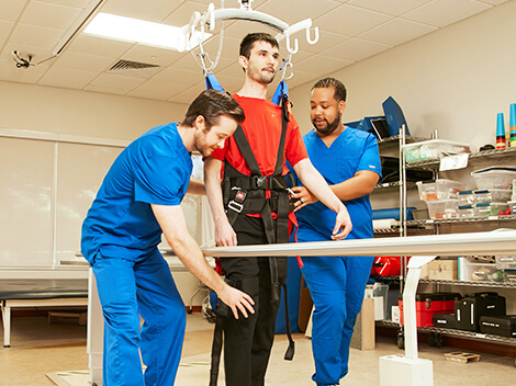 Two male therapists work with a spinal cord injury patient to walk again utilizing an overhead ambulatory track.