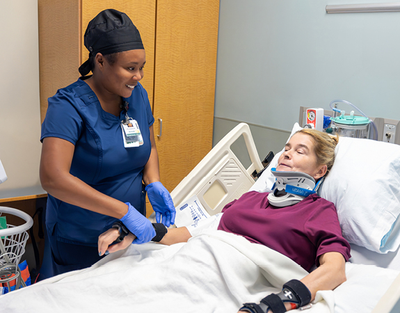 A female patient lays in her hospital bed with her neck stabilized after a spinal cord injury while her nurse takes her vitals.